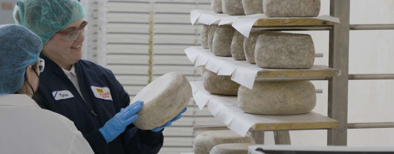 A woman holding a wheel of cheese from a rack of wheels of cheeses